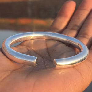 May include: A silver-coloured, open-ended cuff bracelet. The bracelet has a rounded, tubular design and a polished, reflective surface. The bracelet is held in a hand, with a blurred background.