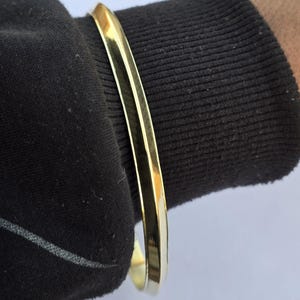 Può includere: Un braccialetto rigido color oro. Il braccialetto ha una superficie liscia e lucida e una forma arrotondata. Il braccialetto è indossato su un polso, in contrasto con il tessuto scuro della manica. Il gioiello è semplice ed elegante.