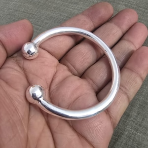 Könnte beinhalten: Ein silberfarbenes Armreif-Armband mit rundem Design. Das Armband hat eine glatte, polierte Oberfläche und an jedem Ende eine kugelförmige Kugel. Der Schmuck wird in einer Hand vor einem neutralen Hintergrund gehalten.