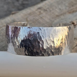 May include: A wide, silver cuff bracelet with a hammered texture. The bracelet has a slightly irregular surface, reflecting light and creating a textured appearance. The cuff is a simple, open design, suitable for everyday wear. The bracelet is approximately 2 inches wide.