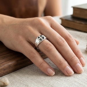 Könnte beinhalten: Ein silberner Siegelring mit einer vertieften kreisförmigen Verzierung, in deren Mitte ein Kreuzsymbol eingraviert ist. Der Ring hat eine polierte metallische Oberfläche und ein breites Banddesign, das schlicht und klar am Finger wirkt.