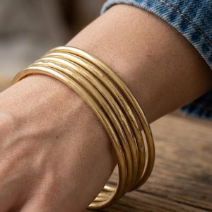 Può includere: Una pila di sei braccialetti color oro su un polso. I braccialetti sono lisci e rotondi, con una finitura leggermente opaca. Il polso è appoggiato su una superficie di legno e sullo sfondo si vede una manica di jeans.
