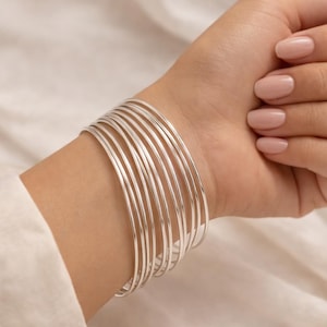 May include: A stack of thin, silver bangles adorns a wrist. The bracelets are smooth and shiny, reflecting light. The skin tone is fair, and the nails are painted a light pink. The background is a soft, neutral color.