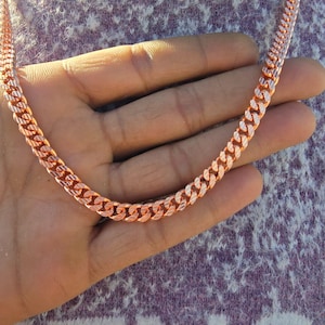 Könnte beinhalten: Eine Halskette mit Panzerkette aus Roségold. Die Kette besteht aus ineinandergreifenden Gliedern, die eine flache, strukturierte Oberfläche bilden. Die Halskette wird auf einer Hand präsentiert, um ihre Farbe und ihr Design zu zeigen. Die Kette hat eine warme, metallische Farbe.
