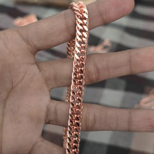 May include: A rose gold chain necklace with a curb link design. The chain is held up against a neutral background. The links are closely connected, creating a smooth, continuous surface. The necklace is a warm, metallic colour.