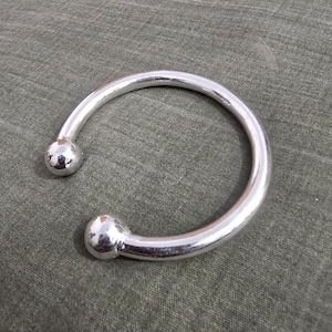 May include: A silver-colored, open cuff bracelet with a rounded design. The bracelet features a curved band that ends in two spherical, polished accents. The jewelry is displayed on a textured, olive-green surface.