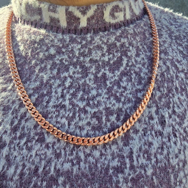 May include: A copper-colored curb chain necklace. The chain is draped over a textured, light purple sweater. The necklace is made of interlocking links, creating a bold and stylish accessory. The sweater has the letters "CHY GIV" on the collar.