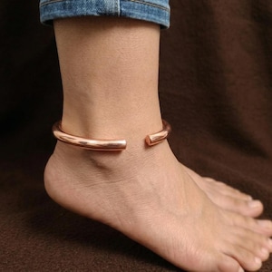 May include: A copper-coloured, open-ended anklet. The smooth, rounded band encircles the ankle, with a small gap at the front. The jewellery is worn on a foot, with a denim-clad leg visible in the background.