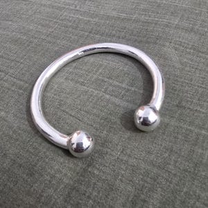 Puede incluir: Un brazalete de puño abierto de color plateado. El brazalete tiene un diseño liso y redondeado con una bola en cada extremo. El metal refleja la luz. El brazalete se muestra sobre una superficie texturizada de color verde oliva.