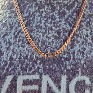 May include: A gold-colored chain necklace with a curb link design. The necklace is displayed against a textured, blue-gray background. The clasp is a simple hook and eye closure.