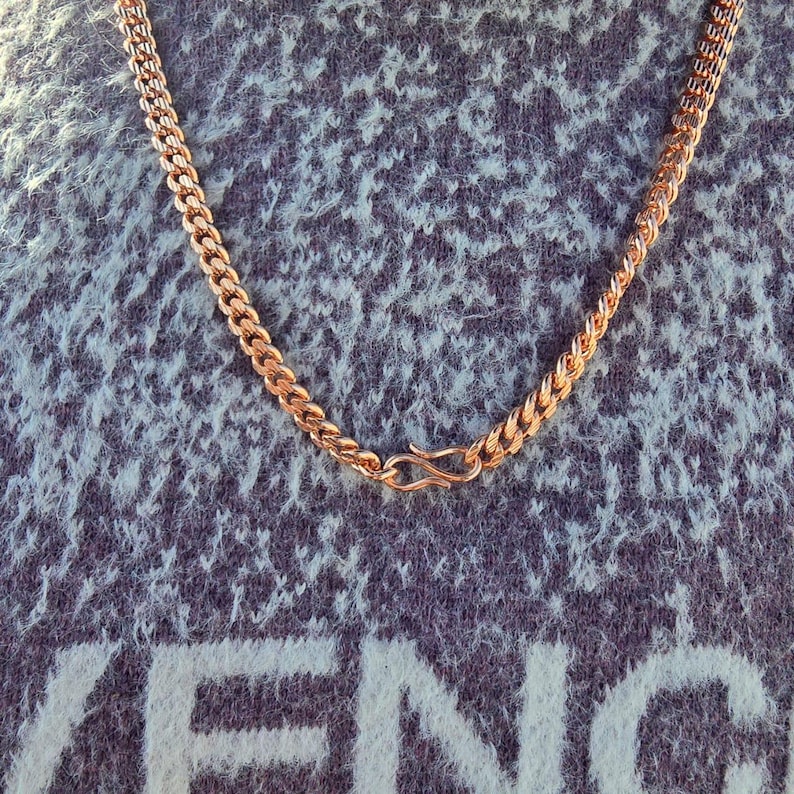 May include: A gold-colored chain necklace with a curb link design. The necklace is displayed against a textured, blue-gray background. The clasp is a simple hook and eye closure.