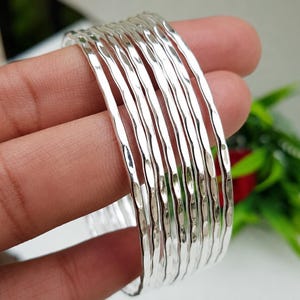 May include: A set of seven individual silver-colored bangles, each featuring a distinct hammered texture that creates a reflective, rippled surface. The bracelets are thin, circular, and designed to be worn together as a stacked set of jewelry.