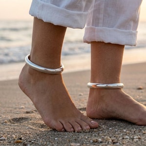 May include: A pair of smooth, polished silver anklets worn on the ankles. The anklets are thick, rounded, and solid in appearance, featuring a reflective metallic finish that catches the light. They are worn above white cuffed trousers.