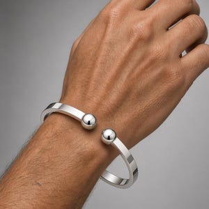 May include: A polished silver-tone cuff bracelet featuring a flat, open-ended band design. The bracelet terminates at both ends with a smooth, spherical ball accent, creating a clean and reflective metallic appearance on the wrist.