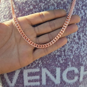 May include: A rose gold chain necklace with a curb link design. The necklace is displayed on a hand, showcasing its color and texture. The chain is made of metal and has a polished finish, reflecting light.