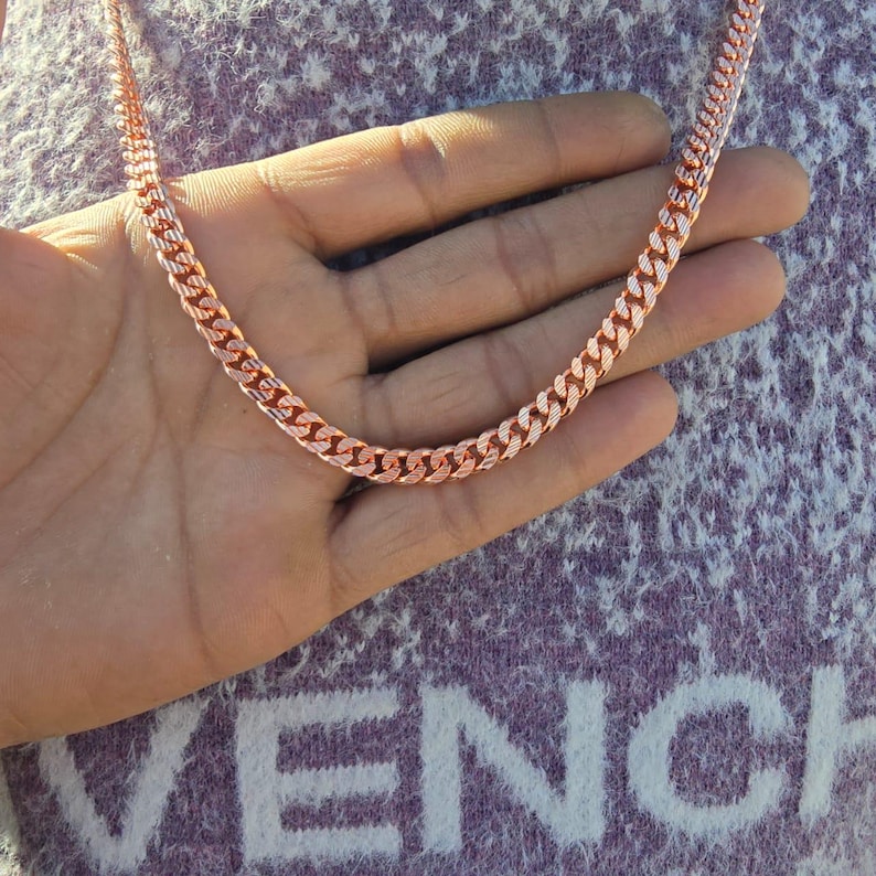 May include: A rose gold chain necklace with a curb link design. The necklace is displayed on a hand, showcasing its color and texture. The chain is made of metal and has a polished finish, reflecting light.