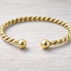 May include: A gold-colored open cuff bracelet with a twisted rope design. The bracelet features a smooth, spherical ball at each end. The jewelry is displayed on a light-colored, textured surface.