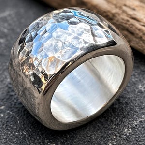 Puede incluir: Un anillo de plata con textura martillada. El anillo tiene una banda ancha y una forma cuadrada redondeada. La superficie refleja la luz, creando una apariencia texturizada. Es una pieza de joyería.
