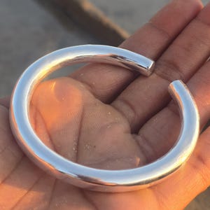May include: A polished silver cuff bracelet with a rounded design. The open-ended bracelet is smooth and reflective, showcasing a simple, elegant style. The bracelet is approximately 6.35 cm in diameter.