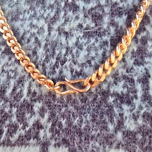 May include: A close-up of a gold-colored chain necklace with a textured, woven design. The necklace features a hook clasp for closure. The chain is displayed against a textured, gray and white background.
