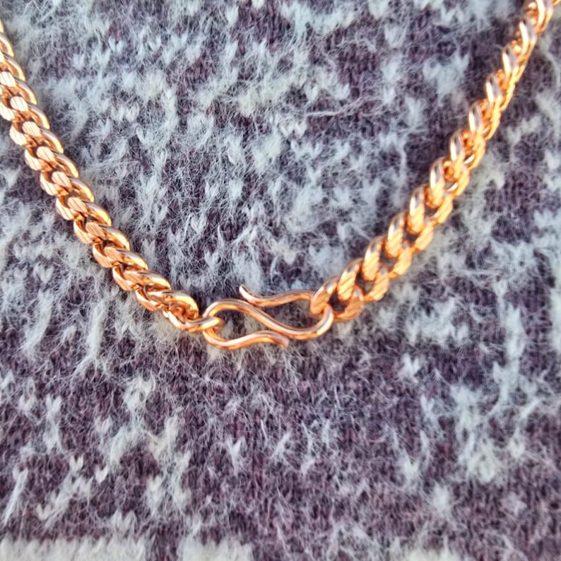 May include: A close-up of a gold-colored chain necklace with a textured, woven design. The necklace features a hook clasp for closure. The chain is displayed against a textured, gray and white background.