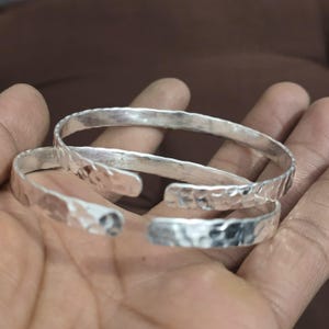 May include: Two silver-colored, hammered-texture cuff bracelets. The bracelets are open-ended and appear to be made of metal. The bracelets are shown on a hand, with the background out of focus.