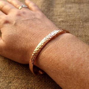 May include: A hammered copper bracelet with a textured surface. The bracelet is a solid band of copper, with small, evenly spaced indentations. The bracelet is worn on a wrist.