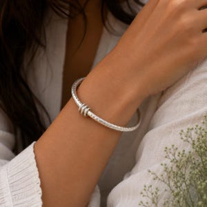 May include: A silver bangle bracelet featuring a textured, hammered surface and three small, smooth, movable silver rings encircling the main band. The bracelet has a circular shape and a metallic finish, reflecting light across its detailed surface.