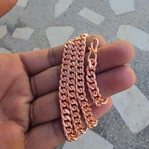 May include: A copper-colored chain necklace with a curb link design. The necklace is displayed on a hand, showcasing the intricate pattern and the warm, metallic hue. The chain has a lobster clasp closure.