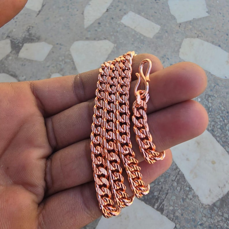 May include: A copper-colored chain necklace with a curb link design. The necklace is displayed on a hand, showcasing the intricate pattern and the warm, metallic hue. The chain has a lobster clasp closure.