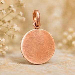 Op de afbeelding: Een ronde hanger van roségoud met een geborstelde, matte afwerking op het platte oppervlak. De hanger is voorzien van een klein, geïntegreerd rond oogje aan de bovenkant voor bevestiging, met een eenvoudig ontwerp in een warme koperkleur.