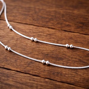Puede incluir: Un delicado collar de plata con pequeñas cuentas redondas espaciadas uniformemente en una fina cadena. El collar se muestra sobre una superficie de madera marrón oscuro, creando un contraste que resalta los detalles de la joya.