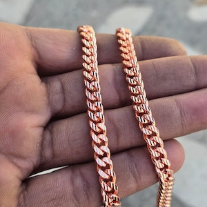 May include: Close-up of two rose gold curb chains. The chains are made of interlocking links, creating a textured surface. The chains are held in a hand, with the links reflecting light.