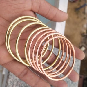 Puede incluir: Un conjunto de brazaletes circulares apilados que presentan una mezcla de acabados metálicos pulidos en tonos dorados y cobrizos. Las pulseras son finas, lisas y de forma redonda, dispuestas en una fila en cascada para mostrar los colores metálicos.