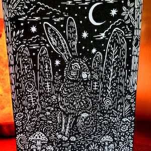 May include: Black and white art print featuring a detailed illustration of a hare in a mystical forest setting. The artwork includes a crescent moon, stars, stylized trees, and mushrooms. The hare is surrounded by standing stones and floral elements.