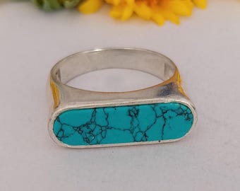 Peut inclure: Une bague en argent avec une incrustation ovale horizontale et allongée en pierre turquoise vive aux veines sombres. La bague est placée devant trois tournesols jaunes, mettant en valeur le contraste entre le métal poli et la pierre.