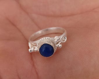 Peut inclure: Une bague en argent ornée d'une pierre précieuse cabochon bleu profond, sertie dans une monture texturée. L'anneau présente un travail métallique complexe avec des détails décoratifs en volutes de chaque côté de la pierre centrale.