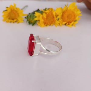 Peut inclure: Une bague en argent avec une grande pierre pr&eacute;cieuse rouge rectangulaire &agrave; facettes mont&eacute;e sur un sertissage &agrave; griffes. L'anneau est lisse et m&eacute;tallique, pr&eacute;sentant une tige fendue. Trois tournesols jaune vif sont en arri&egrave;re-plan.