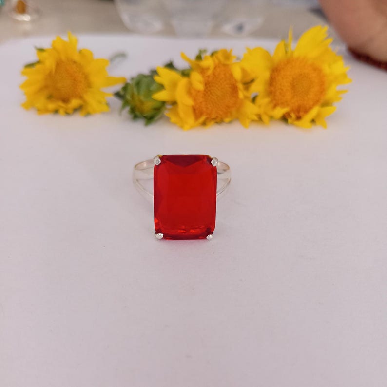 Peut inclure: Une bague en argent avec une grande pierre pr&eacute;cieuse rouge rectangulaire &agrave; facettes, maintenue par quatre griffes. La bague est plac&eacute;e devant trois tournesols jaune vif avec des tiges et des feuilles vertes sur un fond blanc uni.