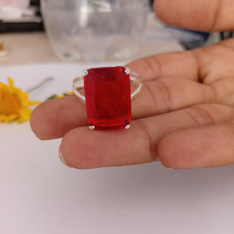 Peut inclure: Une bague en argent avec une grande pierre pr&eacute;cieuse rouge rectangulaire &agrave; facettes, fix&eacute;e par quatre griffes. L'anneau est en m&eacute;tal argent&eacute; poli et pr&eacute;sente un design divis&eacute; qui se connecte &agrave; la base du sertissage de la pierre centrale.