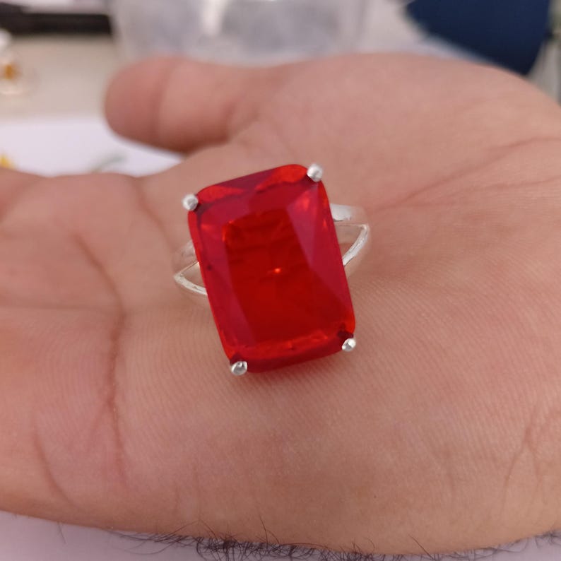 Peut inclure: Une bague en argent avec une grande pierre pr&eacute;cieuse rouge rectangulaire &agrave; facettes, maintenue par quatre griffes m&eacute;talliques. L'anneau est lisse et m&eacute;tallique, mettant en valeur une pierre rouge vive et claire au centre du design.