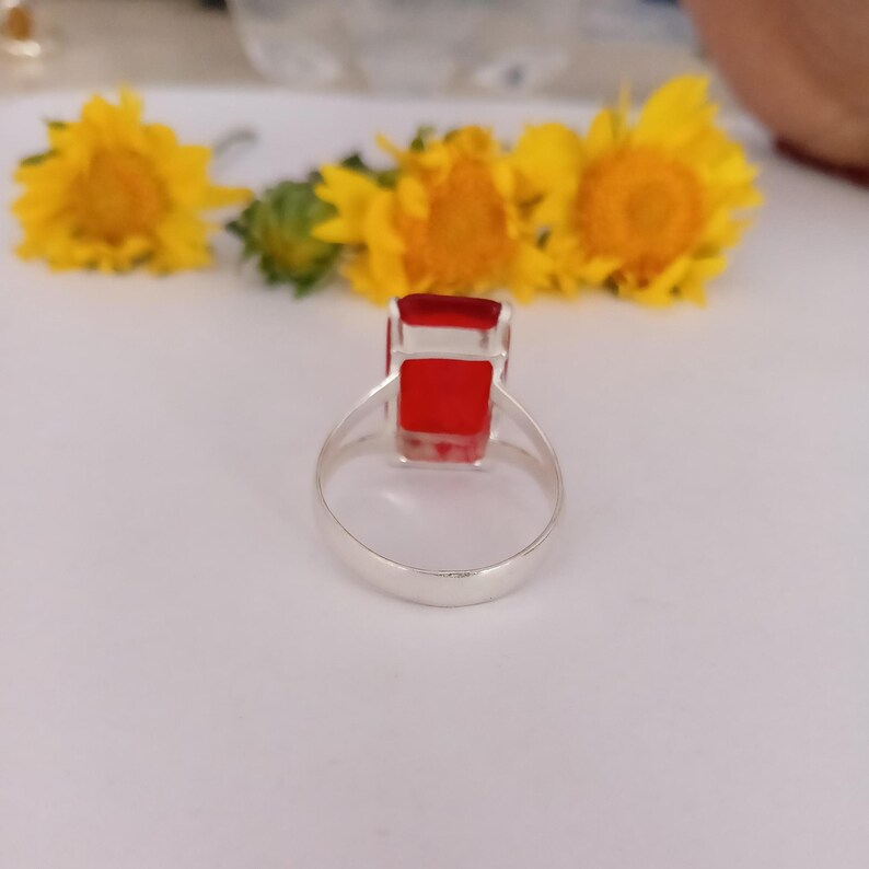 Peut inclure: Une bague en argent avec une pierre pr&eacute;cieuse rectangulaire d'un rouge &eacute;clatant mont&eacute;e sur un anneau m&eacute;tallique simple. La bague est plac&eacute;e verticalement, mettant en valeur la surface claire et polie de la pierre et la finition lisse de l'anneau.