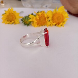 Peut inclure: Une bague en argent avec une pierre pr&eacute;cieuse rouge rectangulaire &agrave; facettes, fix&eacute;e dans une monture m&eacute;tallique &agrave; quatre griffes. L'anneau est lisse et poli, pr&eacute;sentant un design &agrave; tige fendue qui se connecte au sertissage de la pierre.