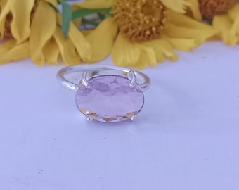Peut inclure: Une bague en argent ornée d'une grosse pierre précieuse ovale rose clair. La bague est mise en valeur sur un fond de fleurs jaunes. La pierre précieuse a une surface à facettes qui reflète la lumière.