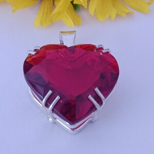 Peut inclure: Pendentif en forme de cœur avec une grosse pierre précieuse rouge rubis facettée. La pierre est sertie dans un cadre en métal argenté. Des fleurs jaunes sont en arrière-plan.