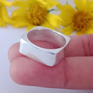 Puede incluir: Un anillo de plata con un diseño cuadrado. El anillo está hecho de un metal brillante y tiene una superficie lisa. El anillo se muestra sobre un fondo de flores amarillas.