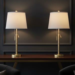 May include: Two matching table lamps with gold-colored bases and off-white shades are displayed on a dark wood table. The lamps are positioned against a dark gray wall, creating a symmetrical and elegant composition.