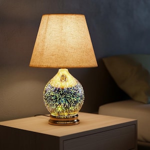May include: A decorative table lamp with a beige fabric shade and a glass base. The base has a spherical shape with a colorful, starburst-like design. The lamp sits on a wooden nightstand in a bedroom setting.