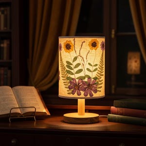 May include: A decorative table lamp with a floral design. The lampshade features sunflowers, purple flowers, and green leaves. The lamp sits on a wooden base, next to an open book and a stack of books. The background includes a bookshelf and a window.
