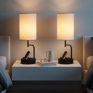 May include: Two matching bedside lamps with cream-colored shades and black bases. Each lamp has a built-in charging station with a phone. A white nightstand holds a mug, glasses, and a book. The lamps are on either side of a bed.